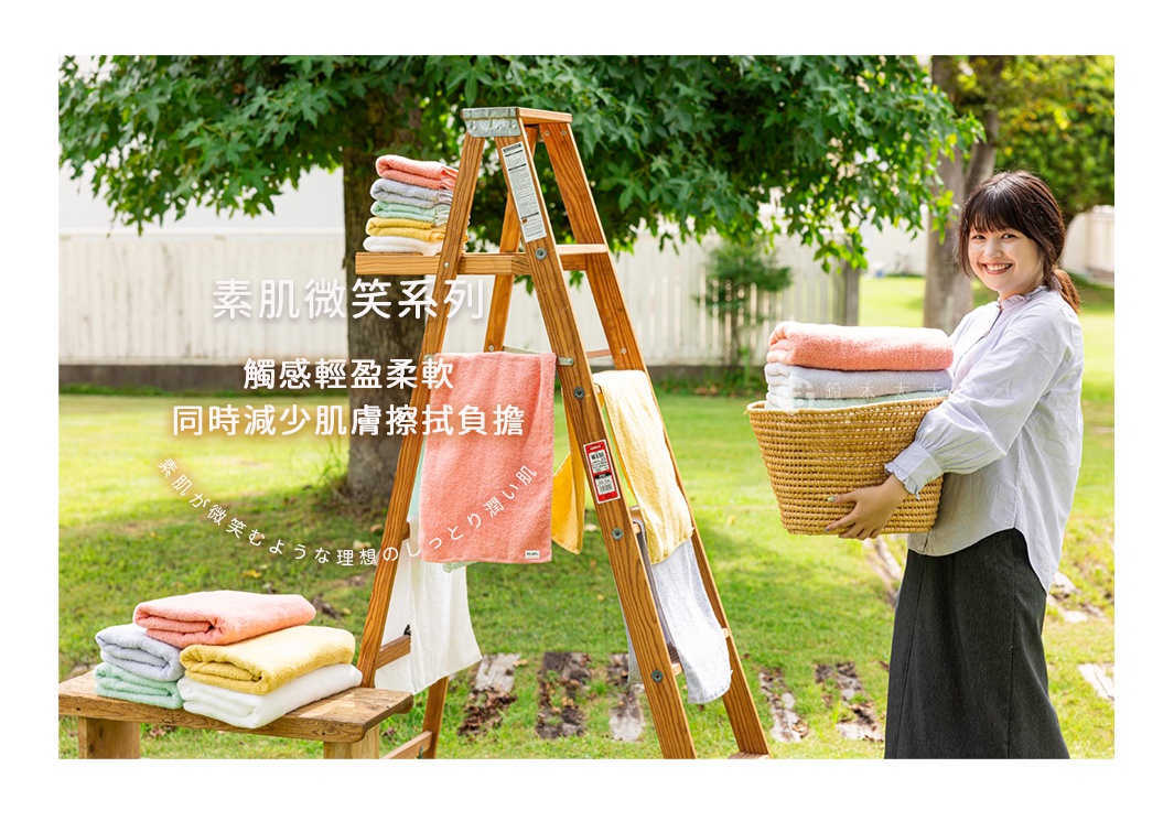 一疊粉色、灰色、淺綠色、黃色及白色毛巾，置於木製梯子和籃子中，旁邊一位女子手提裝有粉色、灰色、淺綠色毛巾的籃子，背景為草地與樹木。