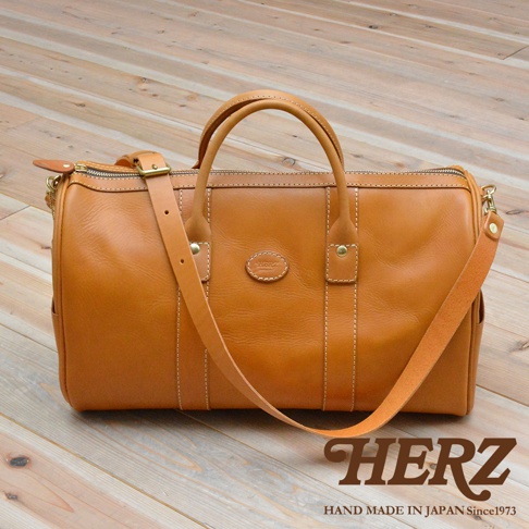 圓筒形棕色皮革肩背手提拉鍊波士頓包，包身縫線清晰，配有可調節肩帶及雙提把，正面中央有一橢圓形皮革標誌，整體質感樸實，風格經典。