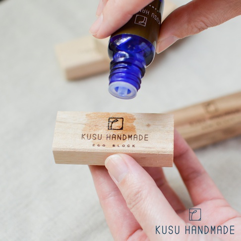 一滴藍色精油滴在刻有「KUSU HANDMADE ECO BLOCK」字樣的淺色木製長方塊上。