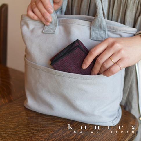 淺灰色帆布托特包，正面有一個較淺的口袋，裡面放著一塊酒紅色、帶有細緻紋理的布料，邊緣有黑色滾邊。