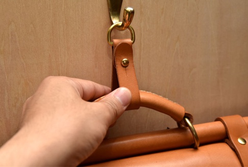 職人手作皮革吊掛扣帶，淺棕色皮革材質，搭配黃銅色金屬環及鉚釘，用於固定和吊掛物品。