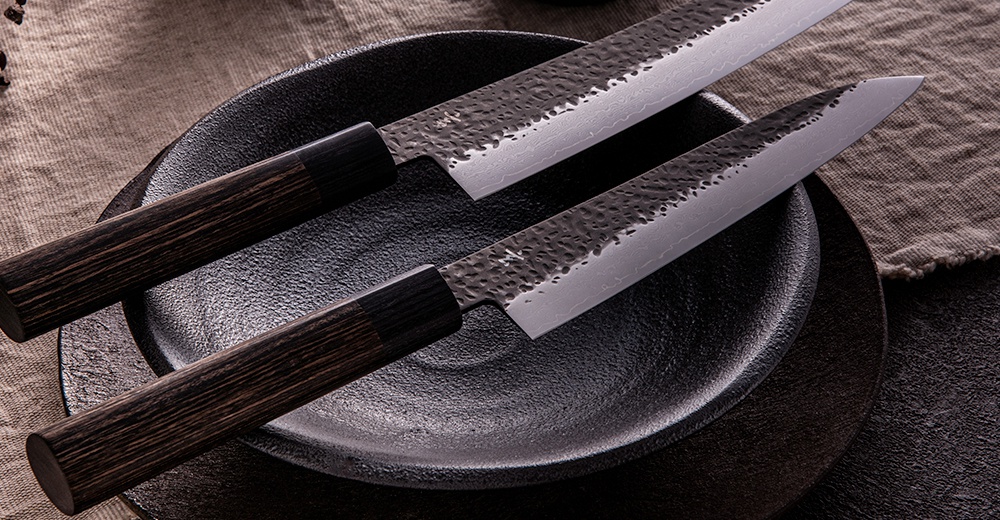 兩把「玄 / Chef's knife 主廚刀21cm」，刀刃為銀色鋼材，帶有槌紋和 Damascus 紋理，刀柄為深棕色木質。刀具平放在黑色圓盤上，背景為麻布。