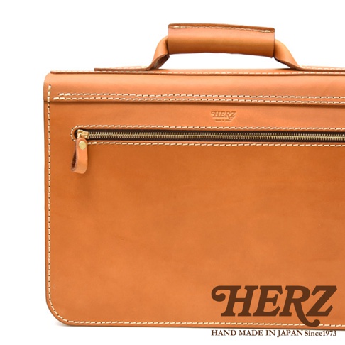 淺棕色皮革製成的手提包，帶有頂部提把、正面拉鍊口袋和白色縫線。