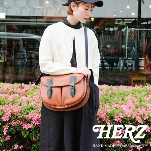 棕色皮革製成的傳統釣魚包，配有黑色搭扣和肩帶，由一位女性模特兒挎著，背景是粉紅色花叢和商店櫥窗。