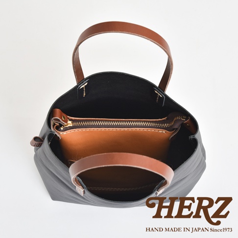 黑色皮革直式隨身單肩包，內部有棕色皮革拉鍊夾層，搭配棕色皮革提把，整體為簡約設計。