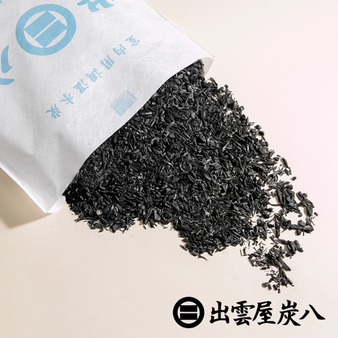 白色紙袋中傾倒出大量黑色、細碎的木炭顆粒，堆積在淺米色背景上。紙袋上有藍色圖案和中文標示「室內調濕木炭」。