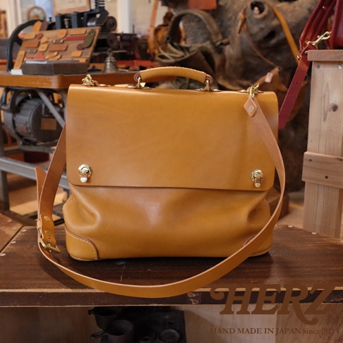 一件棕褐色皮革製作的兩用波士頓包，包身方形，有提把和可拆卸肩帶。包蓋處有兩個金色旋轉鎖扣，邊角處有縫線細節。