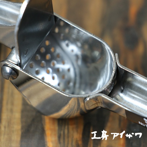 銀色不鏽鋼壓蒜器，帶有多個圓形孔洞的壓榨腔，固定在木質背景上。