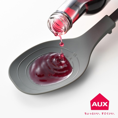 灰色量匙盛著深紅色液體，一瓶玻璃瓶傾斜，液體正從瓶口滴入量匙中。量匙內側有刻度標示。