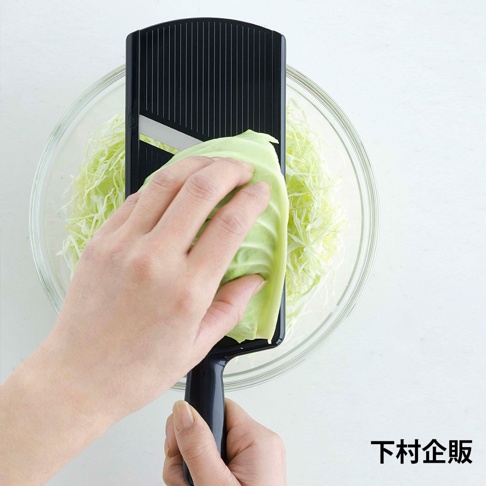 黑色超薄切高麗菜刨絲器，搭配透明玻璃碗中的切絲高麗菜，一隻手正將高麗菜葉置於刨絲器上，另一隻手握住刨絲器的手柄。