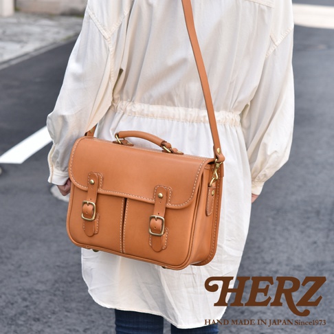 棕褐色皮革兩用單肩包，設有頂部提把、可拆卸肩帶、兩個皮帶扣前袋和黃銅色五金配件。