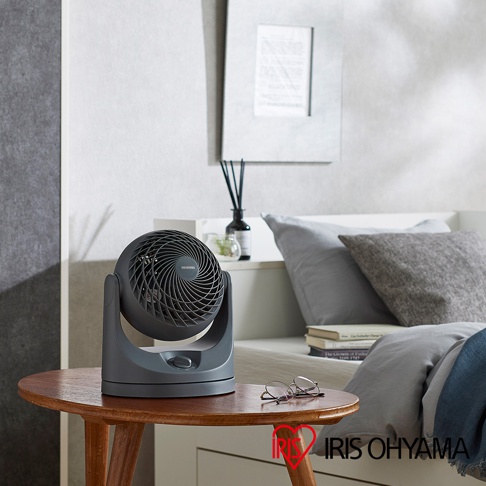 深灰色PCF-MKM15空氣循環扇，帶有可調節的底座，設有旋鈕控制。風扇網罩採用螺旋紋設計，上方印有品牌標誌。產品擺放在木製圓桌上，背景是床鋪和裝飾品。