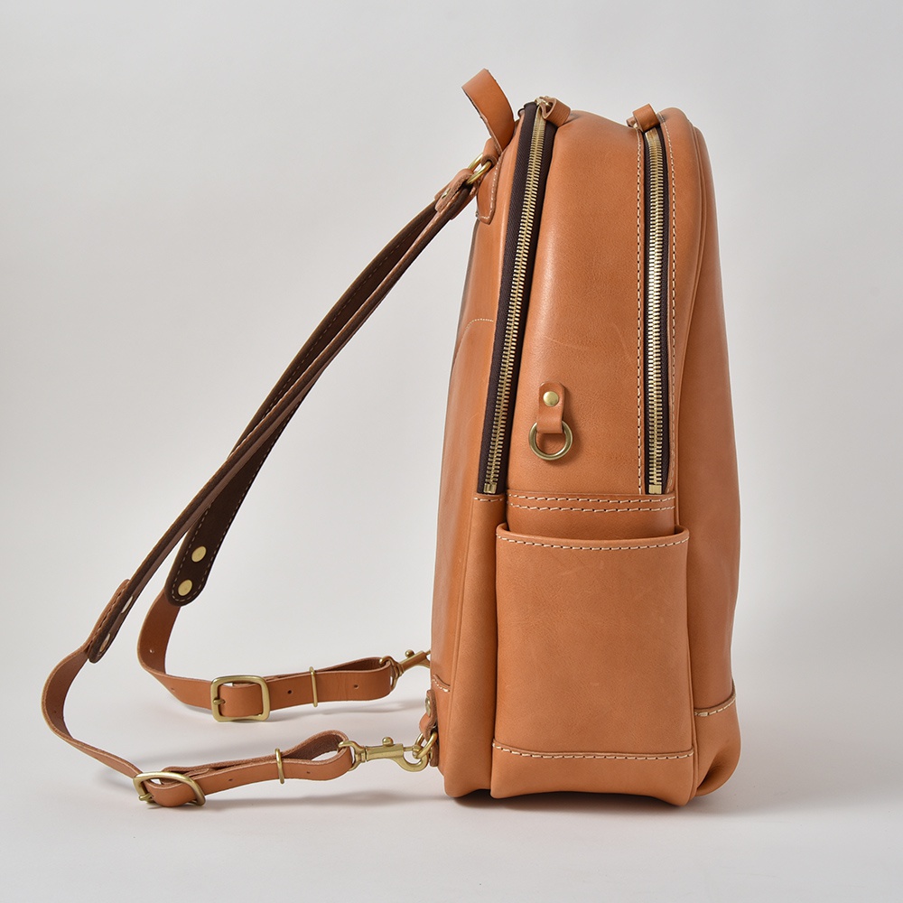 棕褐色皮革多功能後背包，側面有兩條深棕色皮革肩帶，配有黃銅色五金扣環。包身側面有兩個拉鍊隔層，下方有一個開放式口袋，縫線清晰。