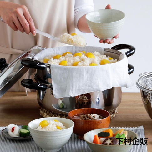 不鏽鋼雙層蒸氣料理鍋，內含蒸籠，蒸籠裡盛滿栗子飯，由白色廚師服的人員用白色飯勺盛飯，旁邊擺放著盛有飯、燉菜和醬菜的日式碗盤。