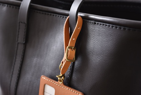 職人手作皮革可調式吊掛帶，附有黃銅扣環，可掛於黑色皮革包上，吊掛帶為淺棕色皮革材質。