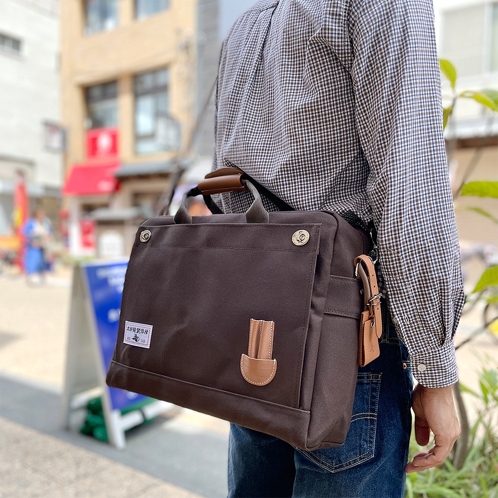 深棕色純綿帆布商務公事包，B4尺寸，配有皮革提把、肩帶扣環及裝飾，左側有品牌布標，整體風格簡潔實用。