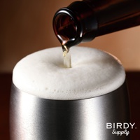 不鏽鋼啤酒杯中，啤酒正從瓶中倒入，形成豐厚綿密的白色泡沫。杯身為銀色不鏽鋼材質，表面帶有細緻的拉絲紋理。