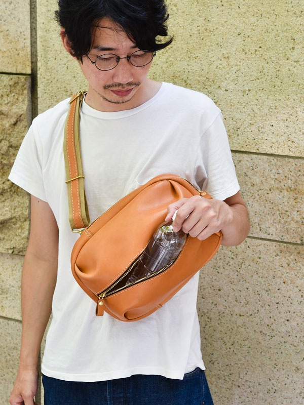 一個男子斜背著一個棕色皮革運動斜背包，包包有一個拉鍊開口，裡面放著一瓶水。