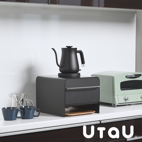 黑色抽屜式收納櫃，頂部放置黑色手沖咖啡壺，櫃體前方有銀色把手，下方露出木質托盤。旁邊有一個淺綠色烤麵包機，櫃子下方是深色木紋廚櫃。