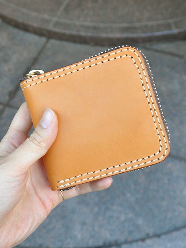 手持一個棕褐色皮革製成的職人手作全包式拉鍊短夾，縫線精緻，拉鍊為銀色，夾身呈現方形，簡約大方。