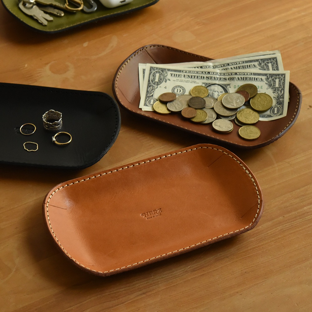 職人手作皮革小物收納盤，共有黑、棕、綠三色。圖片中展示了黑色收納盤內有戒指，棕色收納盤內有美鈔和硬幣，另一個棕色收納盤內有品牌標誌，皮革質感良好，縫線精緻。