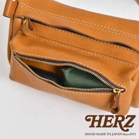 職人手作皮革兩用腰掛斜背包，棕色皮革材質，有拉鍊開合的主袋和前置拉鍊口袋，可見內有綠色物品。