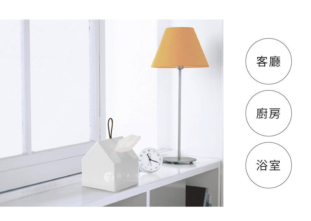白色小屋造型衛生紙盒，頂部有一個黑色掛繩，露出部分白色衛生紙。旁邊有一個銀色底座的橘色燈罩檯燈和一個白色圓形時鐘。