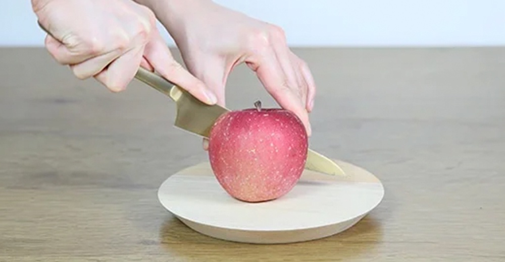 一手持金色水果刀，正在切紅色蘋果，蘋果放在淺色木質圓盤上。