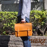 棕色皮革製成的手提公事包，配有兩個金屬釦帶和提把，由穿著牛仔褲和襯衫的人士攜帶，背景為綠色植物和磚牆。