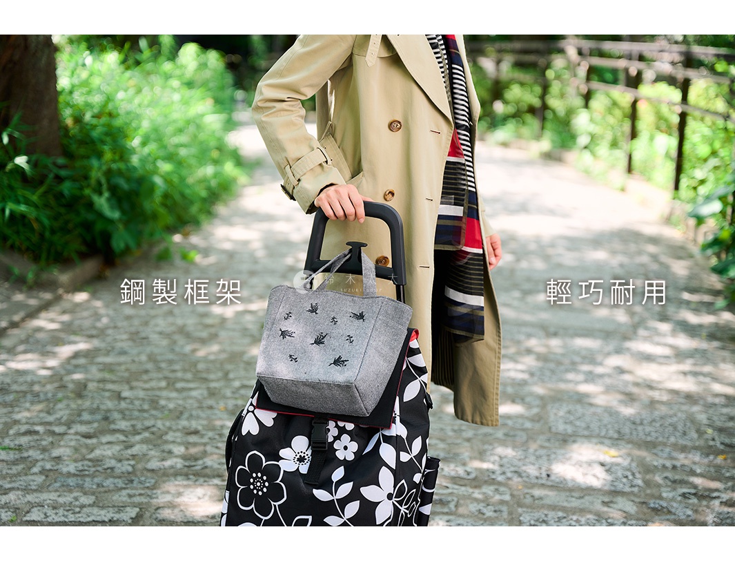 一位身穿卡其色風衣的女士，手提著一個灰色、印有黑色鳥類圖案的折疊購物袋，購物袋下方是一個黑色底、印有白色花朵圖案的購物車。