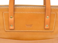 職人手作皮革威靈頓直式拉鍊托特包 (T-151) 的特寫。包身為淺棕色皮革，飾有同色系皮革提把及金色卯釘。正面有壓印的「HERZ」品牌標誌與「MADE IN JAPAN」字樣，周圍以白色縫線勾勒出精緻的口袋輪廓。