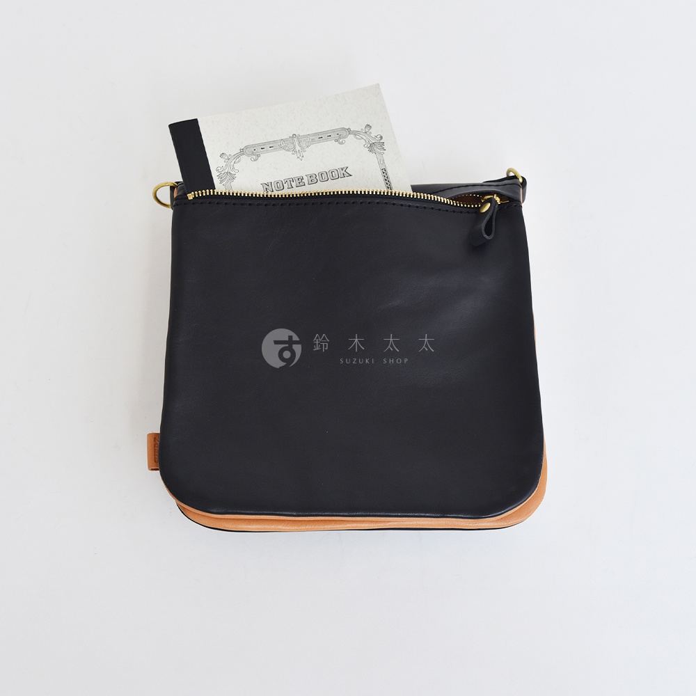 黑色皮革翻轉直式斜背包，底部與側邊為淺棕色皮革，拉鍊開口露出內側的筆記本，包包上有品牌標誌。