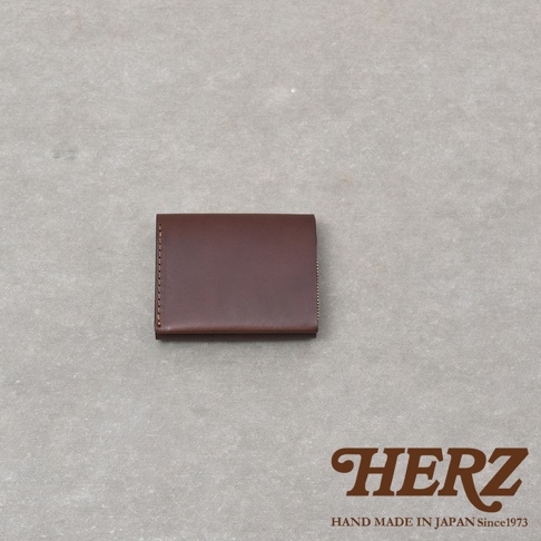 一款放在淺灰色背景上的深棕色日本HERZ手作皮革雙折迷你短夾，呈現簡約俐落的皮革質感，右下角印有HERZ品牌字樣及手作標語。