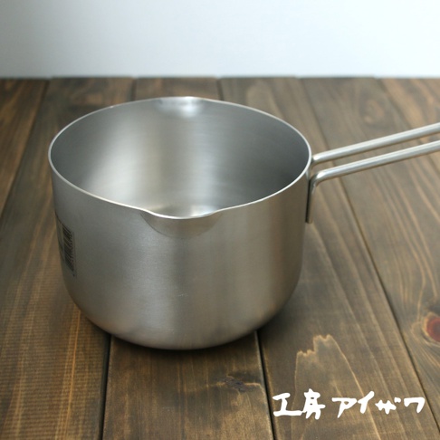 一個14公分的銀色不鏽鋼雙口牛奶鍋，鍋身圓潤，帶有兩個平行銀色把手，放置在木紋桌面上。