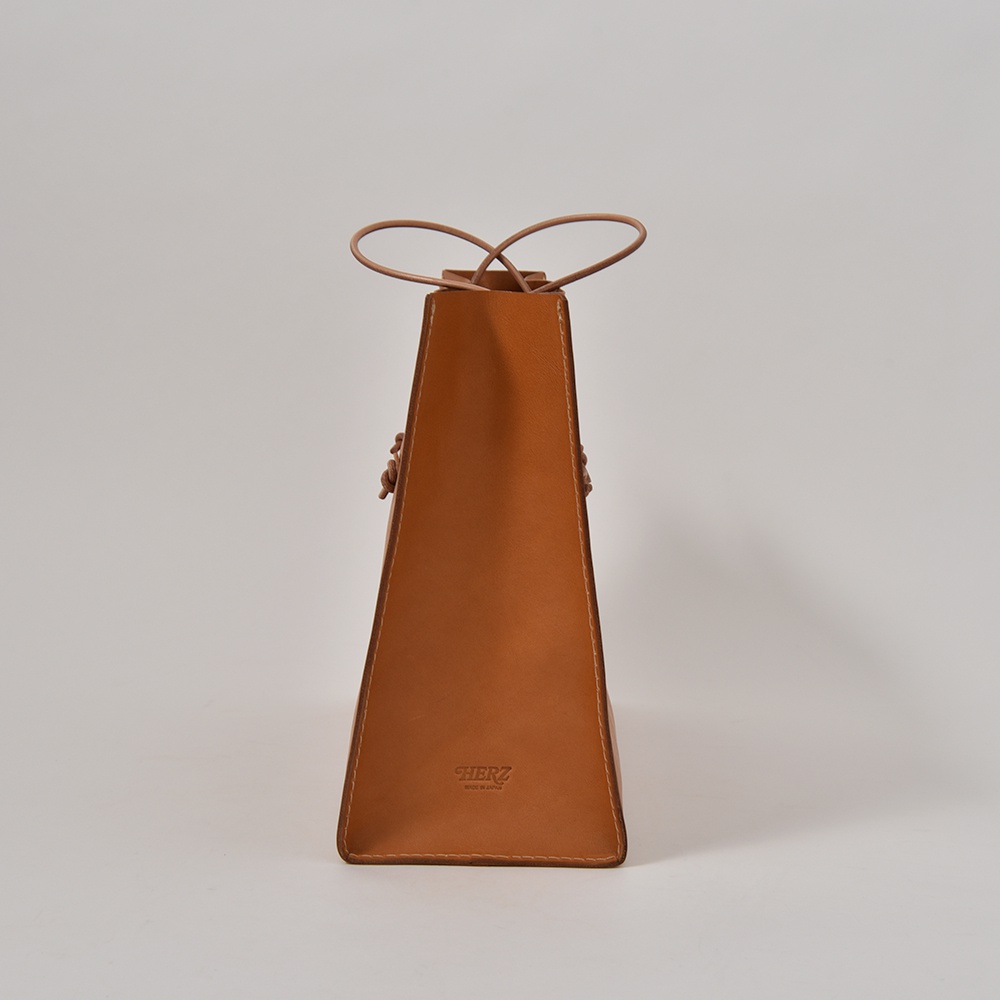 職人手作皮革提袋 T-5，呈現簡潔的梯形輪廓，採用光滑的棕褐色皮革製成，配有兩條細長的皮革提把，於袋口處打結。袋身側面有細緻的縫線，底部印有品牌標誌。