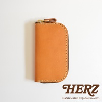 駝色皮革橢圓形拉鍊鑰匙包，配有金色拉鍊及皮革拉環，包身縫線細緻，底部有HERZ品牌標誌，標示HAND MADE IN JAPAN Since1973。