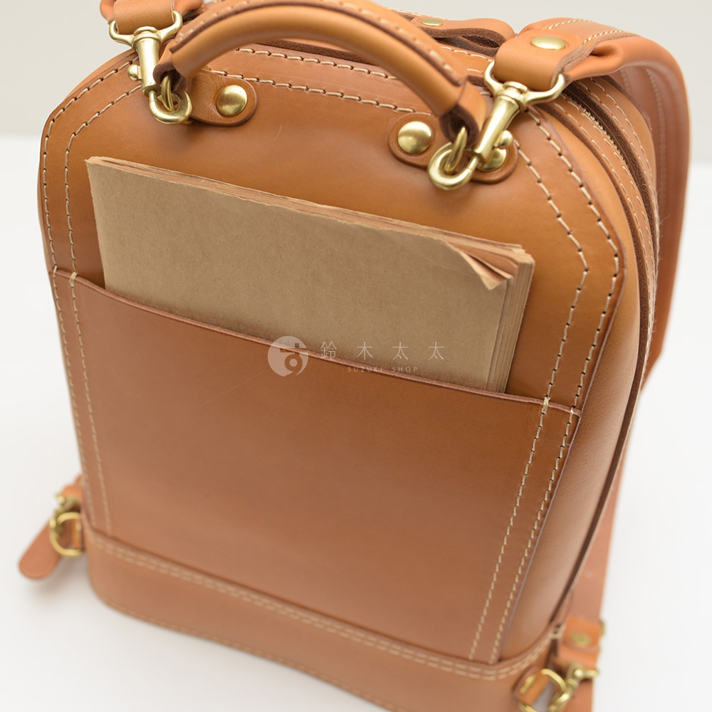 棕色皮革製成的手提後背包，配有金屬扣環和白色車縫線，正面有一個插袋，內有兩本筆記本。