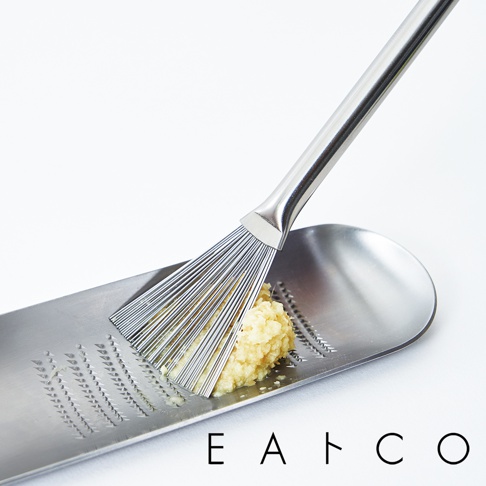 銀色不鏽鋼製的日製料理小刷，刷毛呈扇形展開，正用於刮取長條形不鏽鋼磨泥器上的蒜泥。磨泥器表面有細密的刮紋和集泥槽。