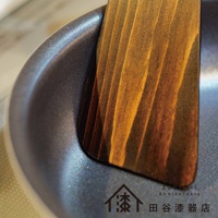 輪島塗漆能登檜料理鏟，木紋清晰，呈現深淺不一的天然木色，鏟面邊緣與灰色鍋具的圓潤曲面緊密貼合。