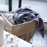 一條淺藍色、柔軟的舒眠雙面四季毯，疊放在一個米色帆布內襯的儲物籃中，旁邊還有另一條淺色毯子。