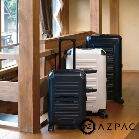 三款 AZPAC Trucker 旅行箱，分別為黑色、白色、黑色，皆為 30 吋尺寸，具備防爆拉鍊和可伸縮拉桿，箱體呈現條紋設計，置於木質地板和樓梯旁。