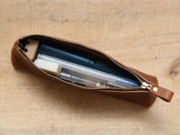 職人手作棕色皮革三角收納筆袋，拉鍊部分為金色，袋內裝有藍色筆、白色筆和橡皮擦。