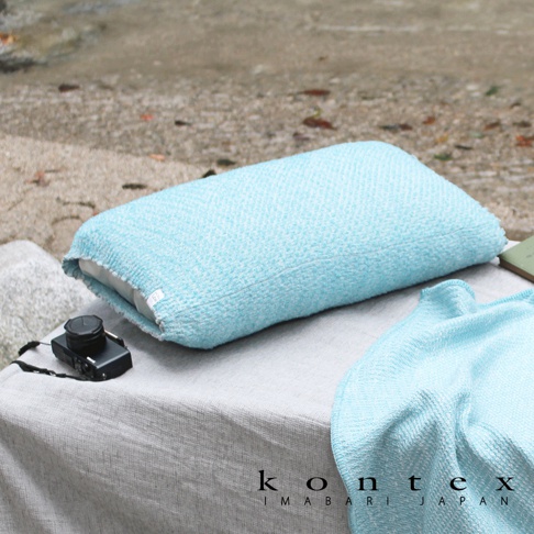 KONTEX IMABARI JAPAN 品牌標誌，以及藍色枕頭和毛巾，擺放在沙灘旁的桌子上，旁邊還有一台小相機。