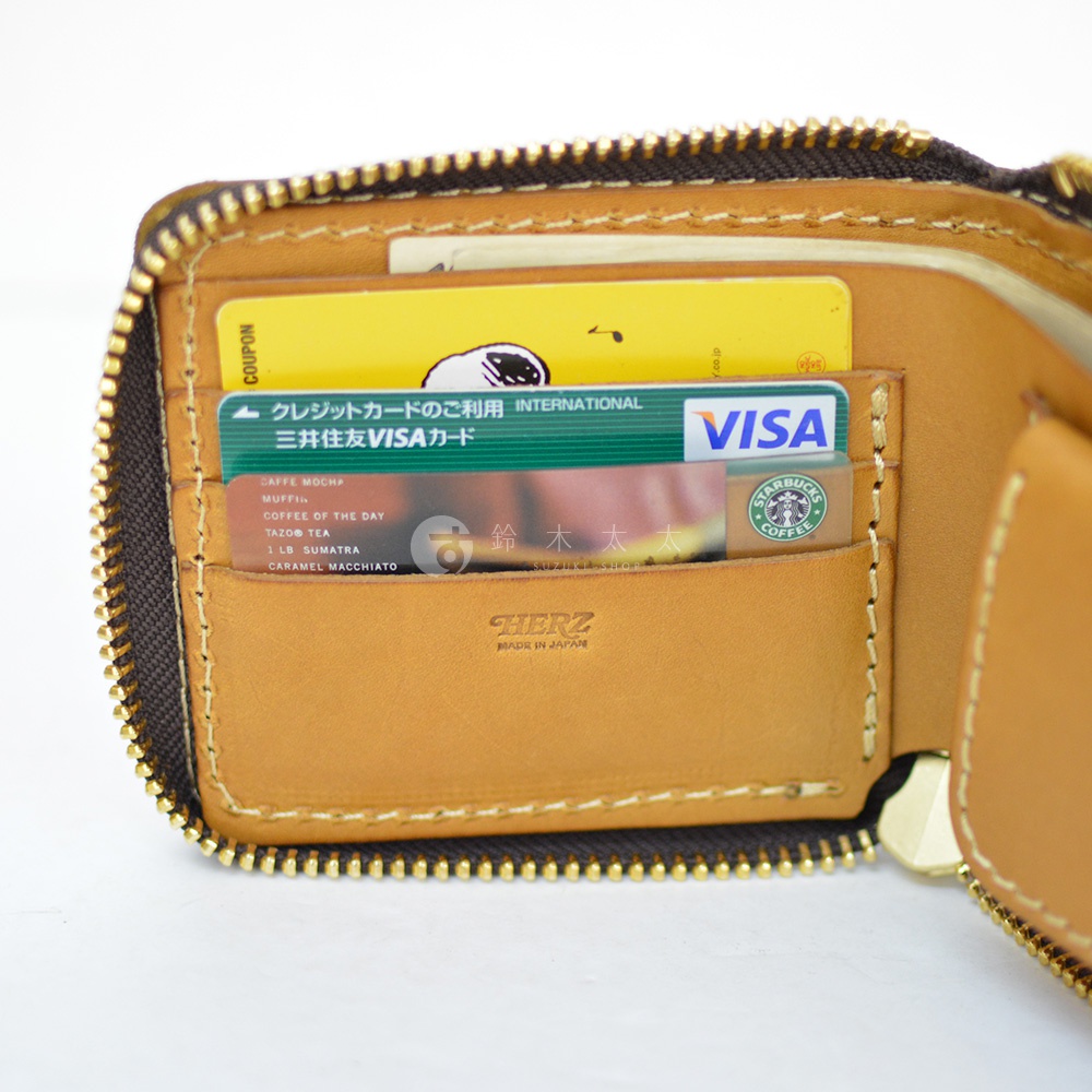 一張日本HERZ職人手作皮革短夾的內部特寫照，棕色皮革質感細膩且縫線工整，拉鍊圍繞邊緣設計。夾層中插有Visa信用卡、星巴克儲值卡及黃色折價券，皮夾內側壓印有HERZ與made in Japan的字樣。