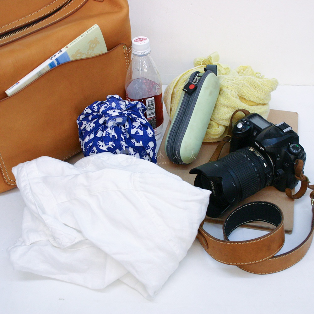 棕褐色皮革厚實單肩波士頓包，內有藍白動物紋束口袋、白色衣物、綠色筆袋、黃色圍巾、水瓶及黑色單眼相機。