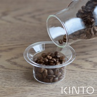 透明玻璃量杯中裝滿咖啡豆，另一玻璃容器傾斜，咖啡豆正倒入量杯。量杯邊緣有「20g」刻度，整體置於木質桌面。