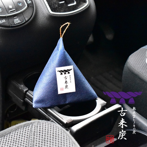 一個藍色三角形的古來炭除濕包放置在汽車杯架中，包裝袋上有帶有注連繩圖案的標籤與奧出雲檜木炭字樣，整體呈現日式簡約風格。
