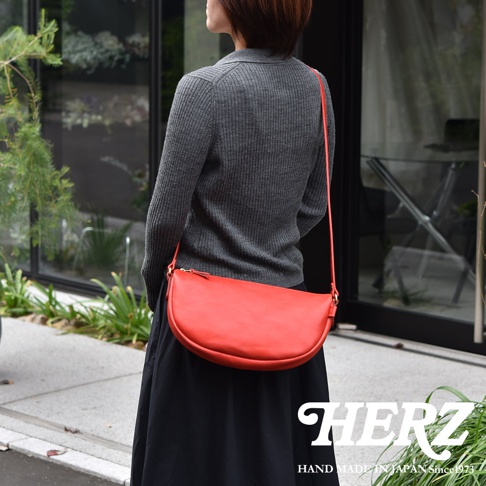 日本HERZ職人手作皮革半圓馬鞍包，紅色彎月形包身，搭配灰色針織衫與黑色長裙。
