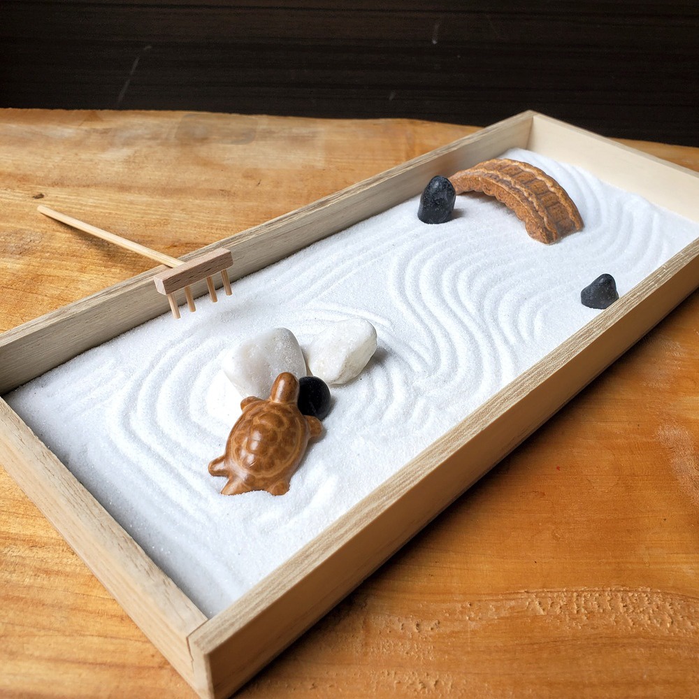 日式枯山水模型，淺木框內鋪滿白色細沙，沙上刻有漣漪紋路。模型包含一隻褐色烏龜雕塑、兩顆白色鵝卵石、一顆黑色鵝卵石、一座褐色石拱橋及一支木柄小耙子。