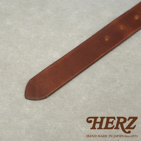 HERZ 品牌標誌與標語 HAND MADE IN JAPAN Since1973，近景呈現一條深棕色皮革腰帶，皮帶上有幾個打孔，背景為淺灰色紋理。
